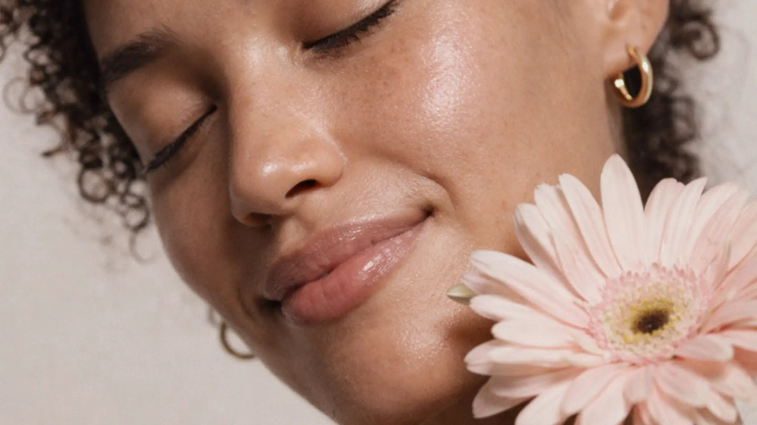Close-up of a woman with glowing skin holding a pink flower near her face.